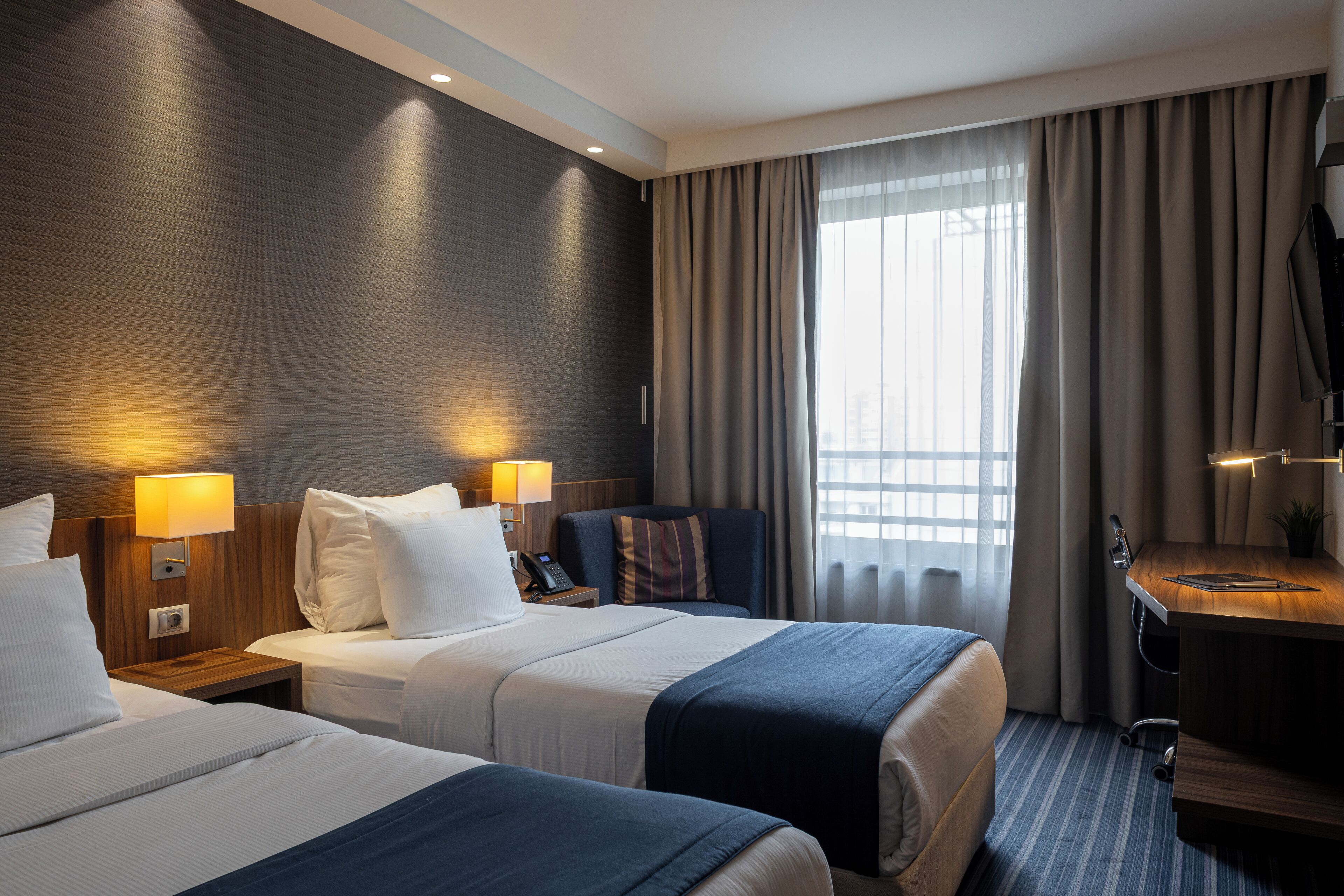 A hotel room with two beds, a desk, a TV, and a window with curtains.