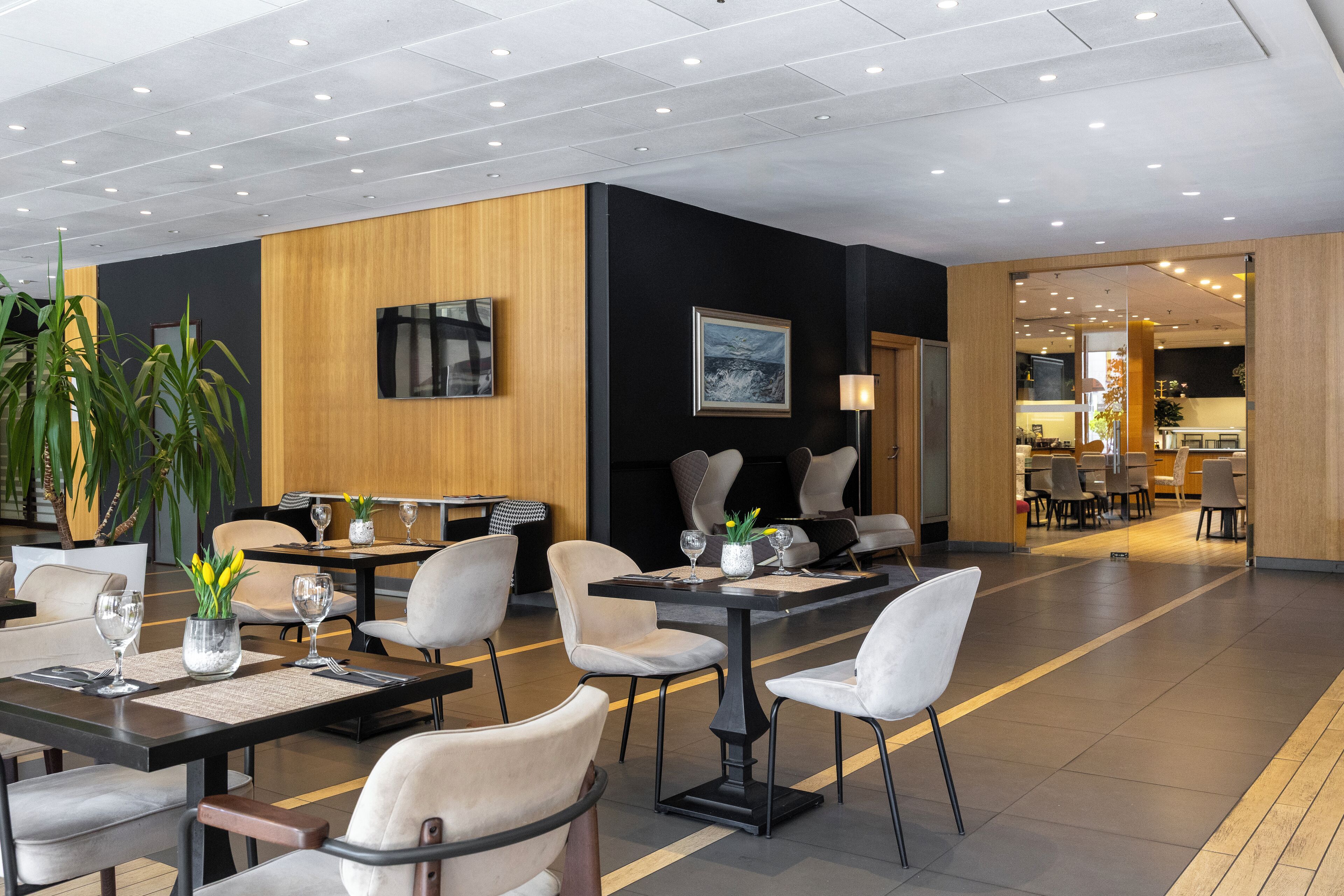 Um hall de hotel moderno com várias mesas e cadeiras para refeições, uma grande planta em vaso e um quadro na parede.