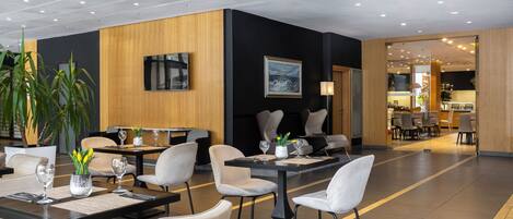Um hall de hotel moderno com várias mesas e cadeiras para refeições, uma grande planta em vaso e um quadro na parede.
