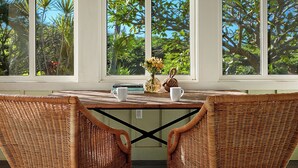 A wicker table with two chairs, a vase with flowers, and books on it, set against a window with a view of tropical trees. - Hale Milo: Charming Garden Cottage in Sunny Poipu! (Koloa)