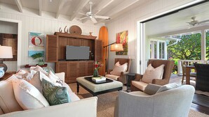 A spacious living room with a large television, comfortable seating, and a view of the outdoors. - Hale Milo: Charming Garden Cottage in Sunny Poipu! (Koloa)