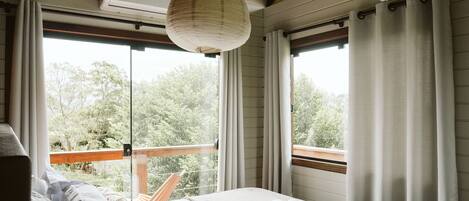 Une chambre à coucher avec un lit, des oreillers, un luminaire suspendu et une vue sur les arbres à travers de grandes fenêtres.