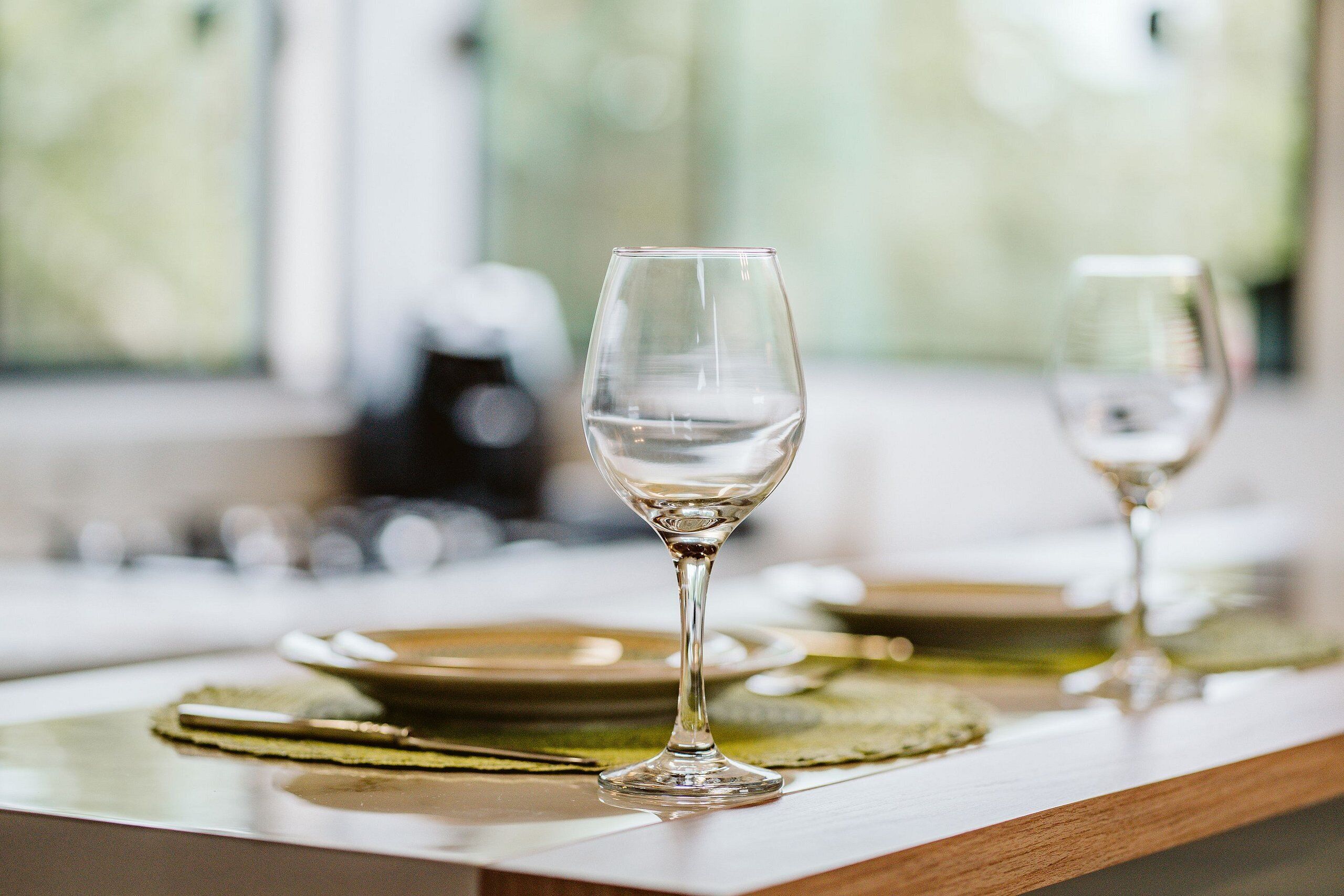 Mesa de jantar posta com dois copos de vinho vazios e pratos, com jogos americanos verdes.
