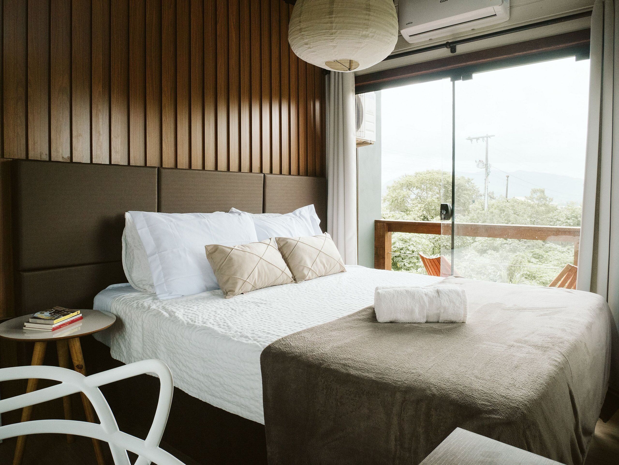 Un dormitorio con una cama grande, un cabecero de madera, una mesita de noche con libros y vistas a la vegetación a través de una ventana.