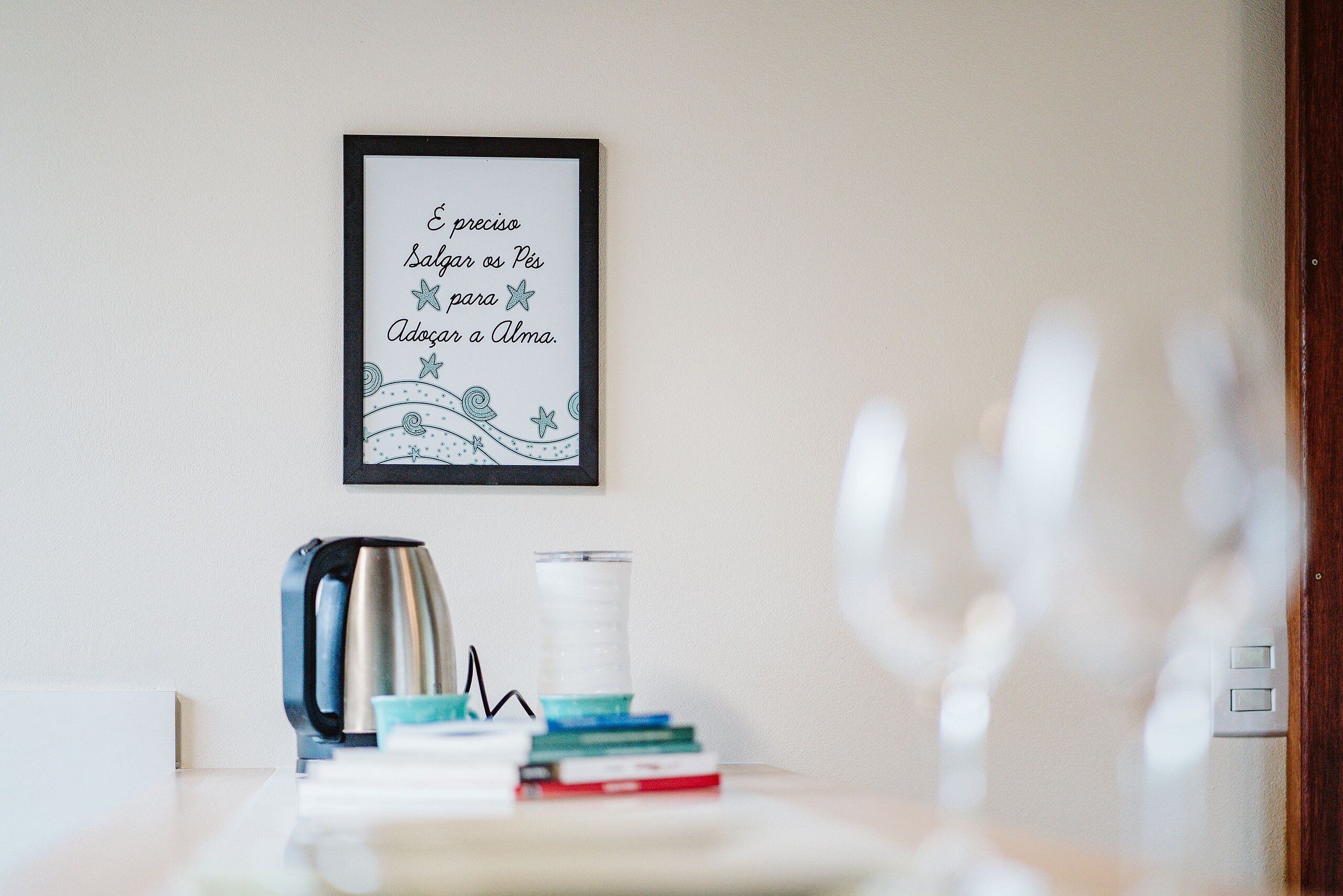 A framed quote on a wall, a kettle, a stack of books, and a glass on a table.