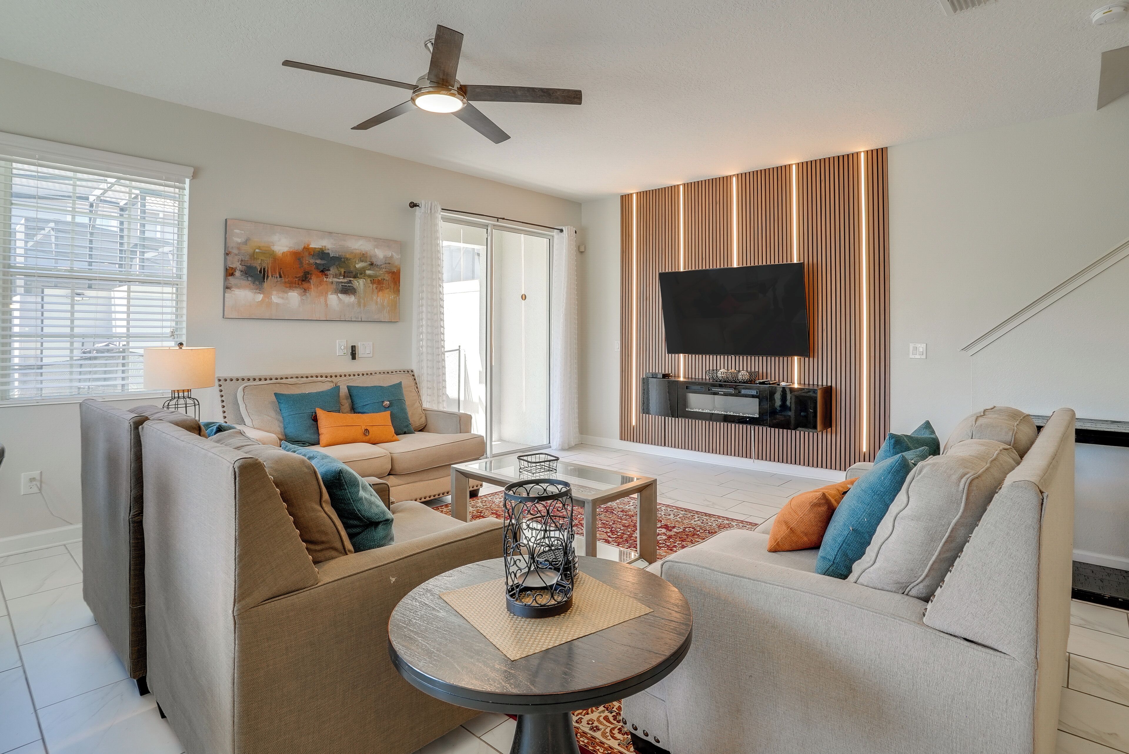 A modern living room with a sofa set, a coffee table, a flat-screen TV mounted on a wooden panel, and a ceiling fan.