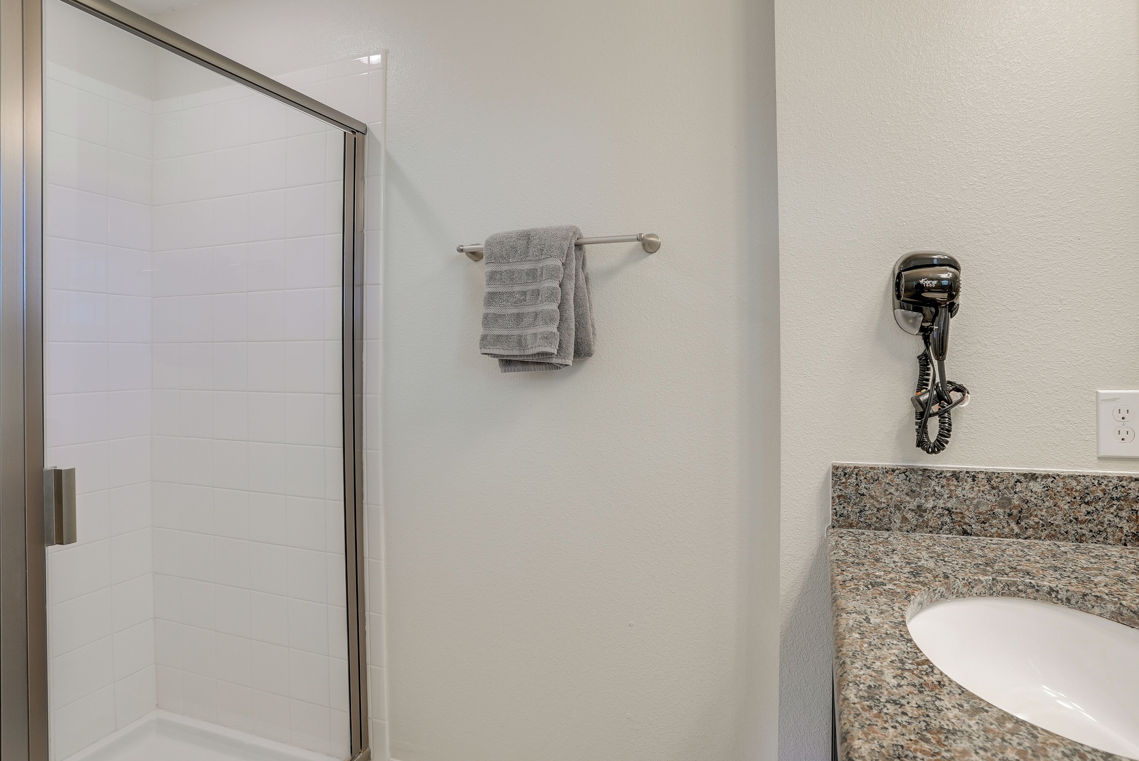 A bathroom with a glass shower door, a towel rack with a folded towel, a hairdryer mounted on the wall, and a granite countertop with a sink.