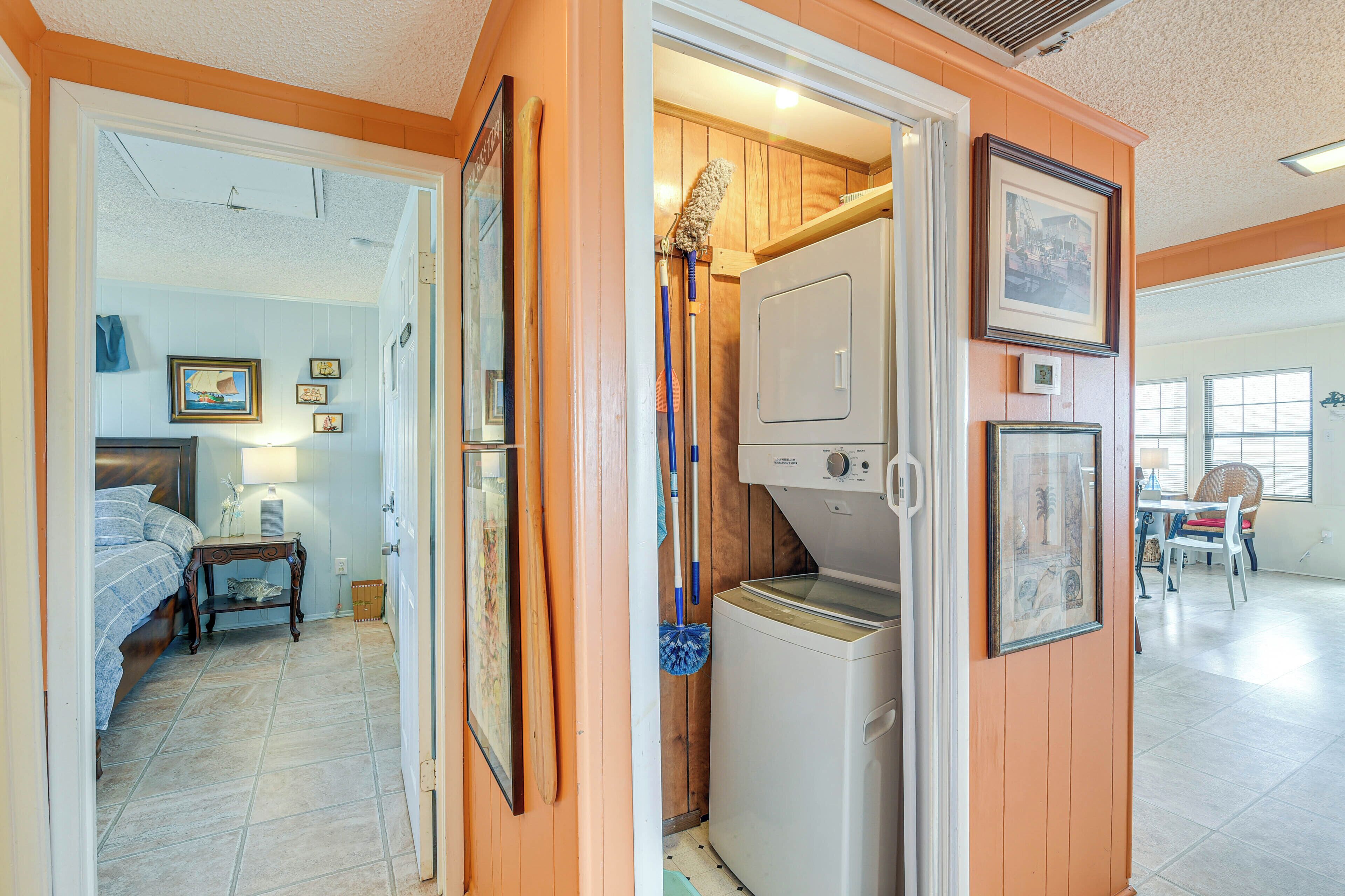 A laundry room with a washing machine, a mop, and a broom. There is a bed and a nightstand with a lamp in the adjacent bedroom.