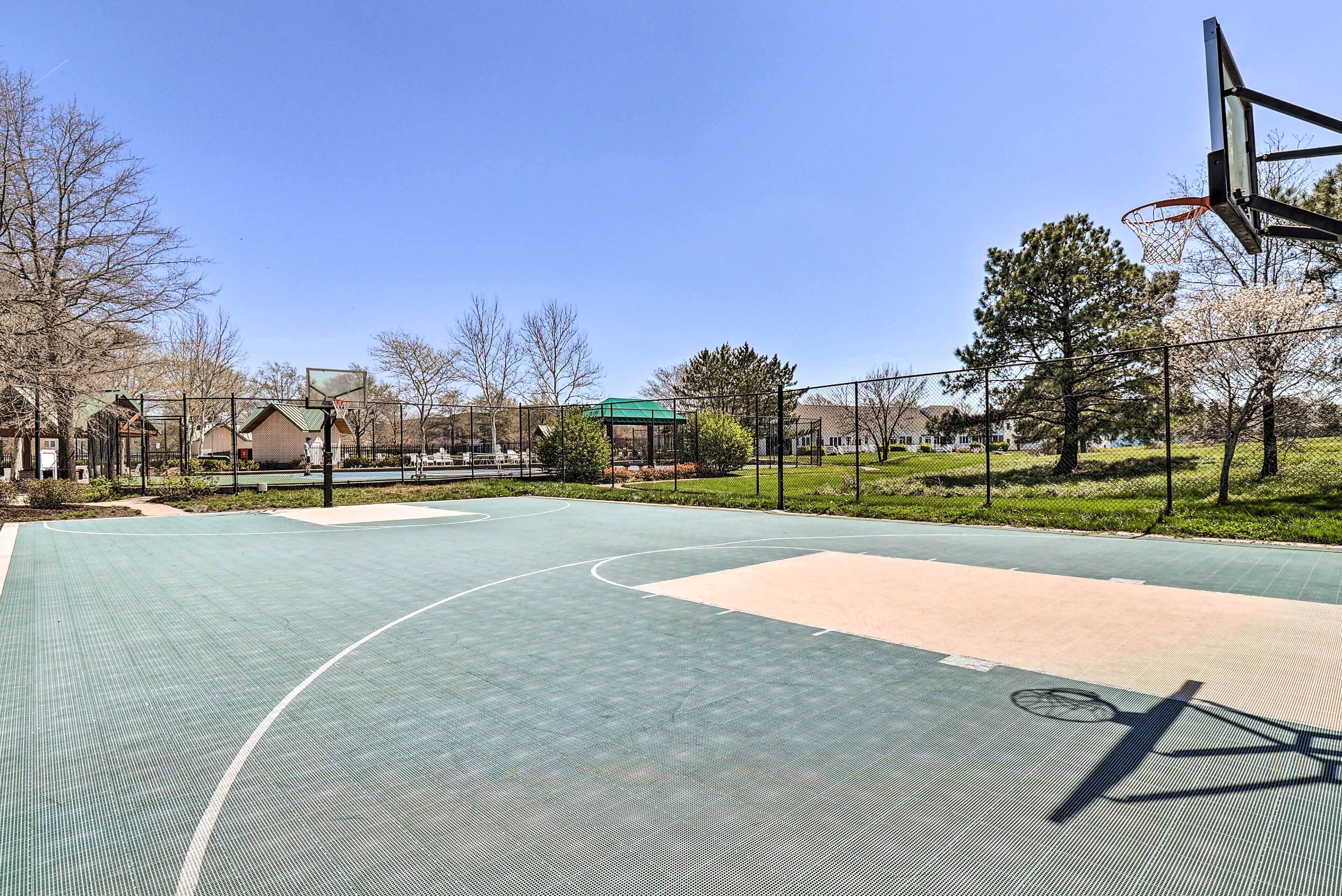 An outdoor basketball court with a green surface, surrounded by a fence, trees, and houses in the background.