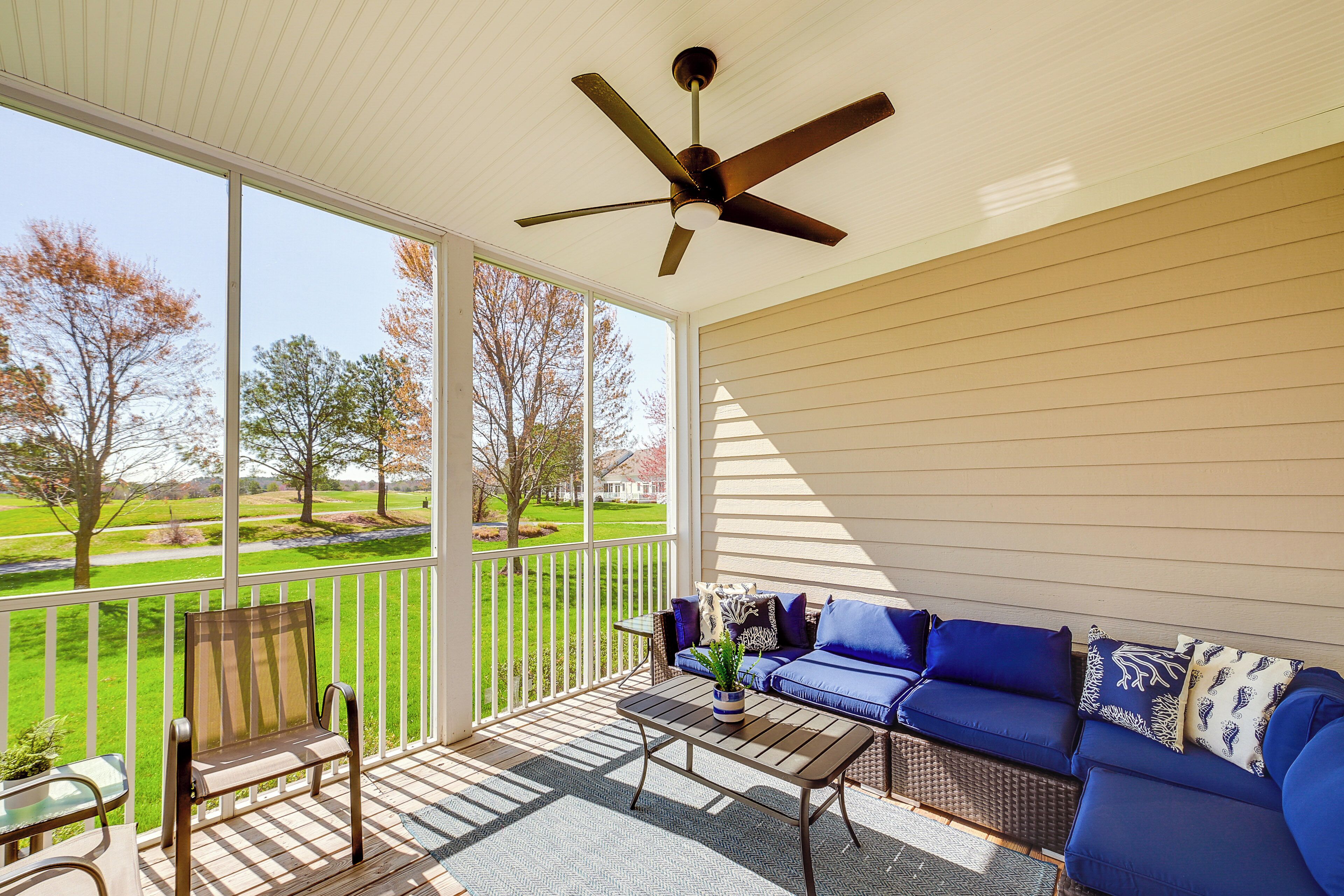 A covered porch with a view of a green field, a blue sofa, and a ceiling fan.