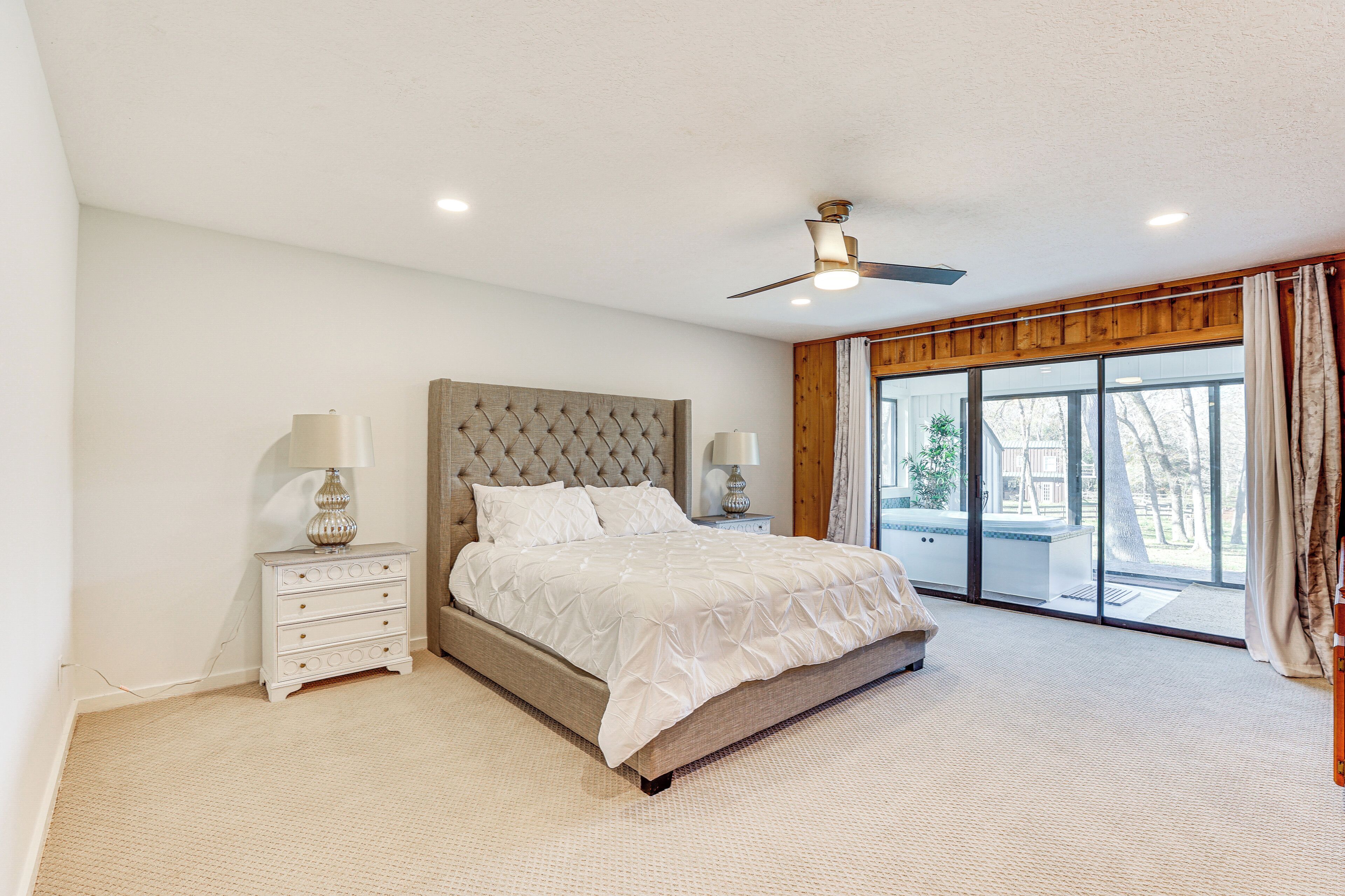 A bedroom with a large bed, two bedside tables, a ceiling fan, and a view through a large window.