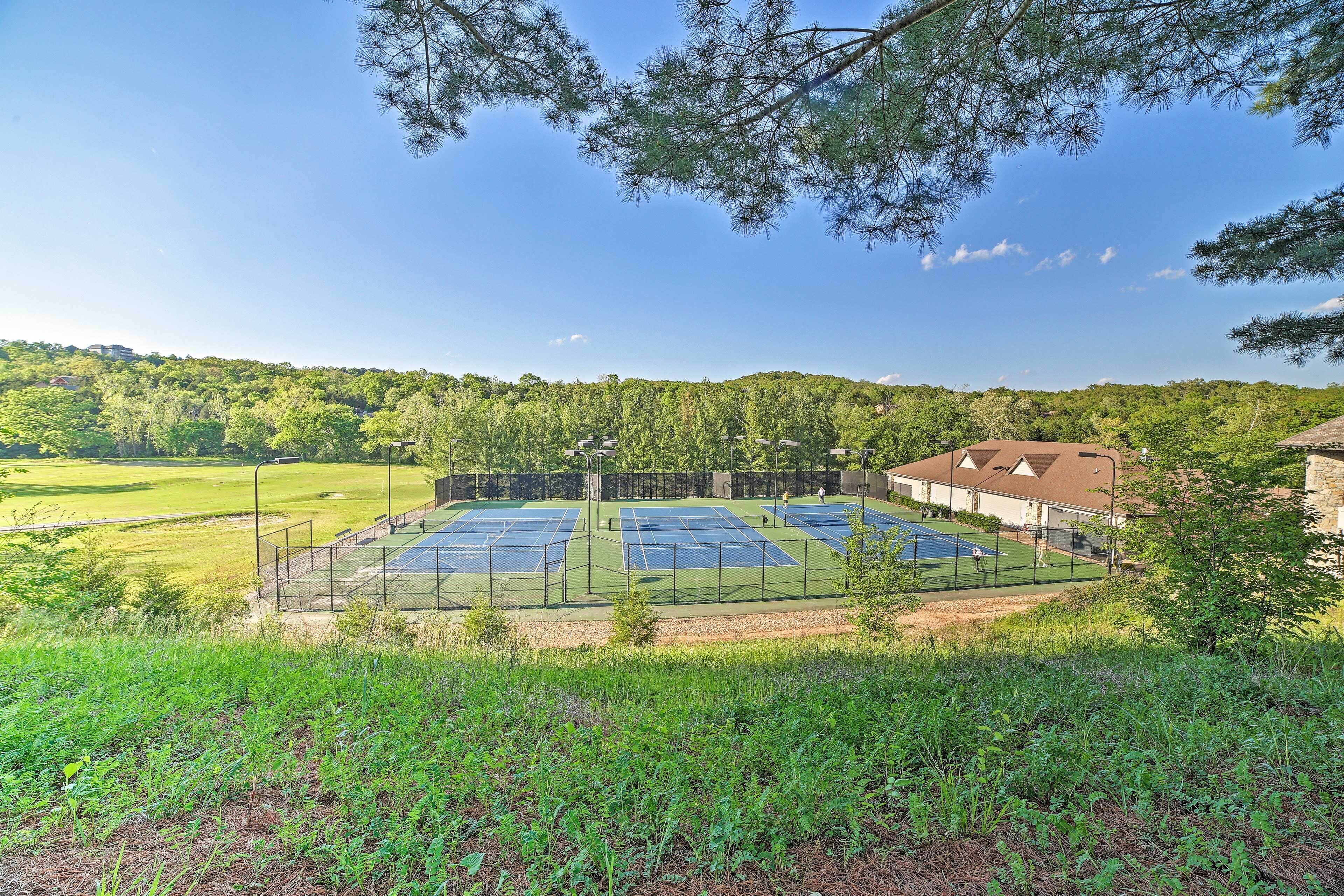 A tennis court complex with multiple courts, surrounded by greenery and a building with a sloped roof.
