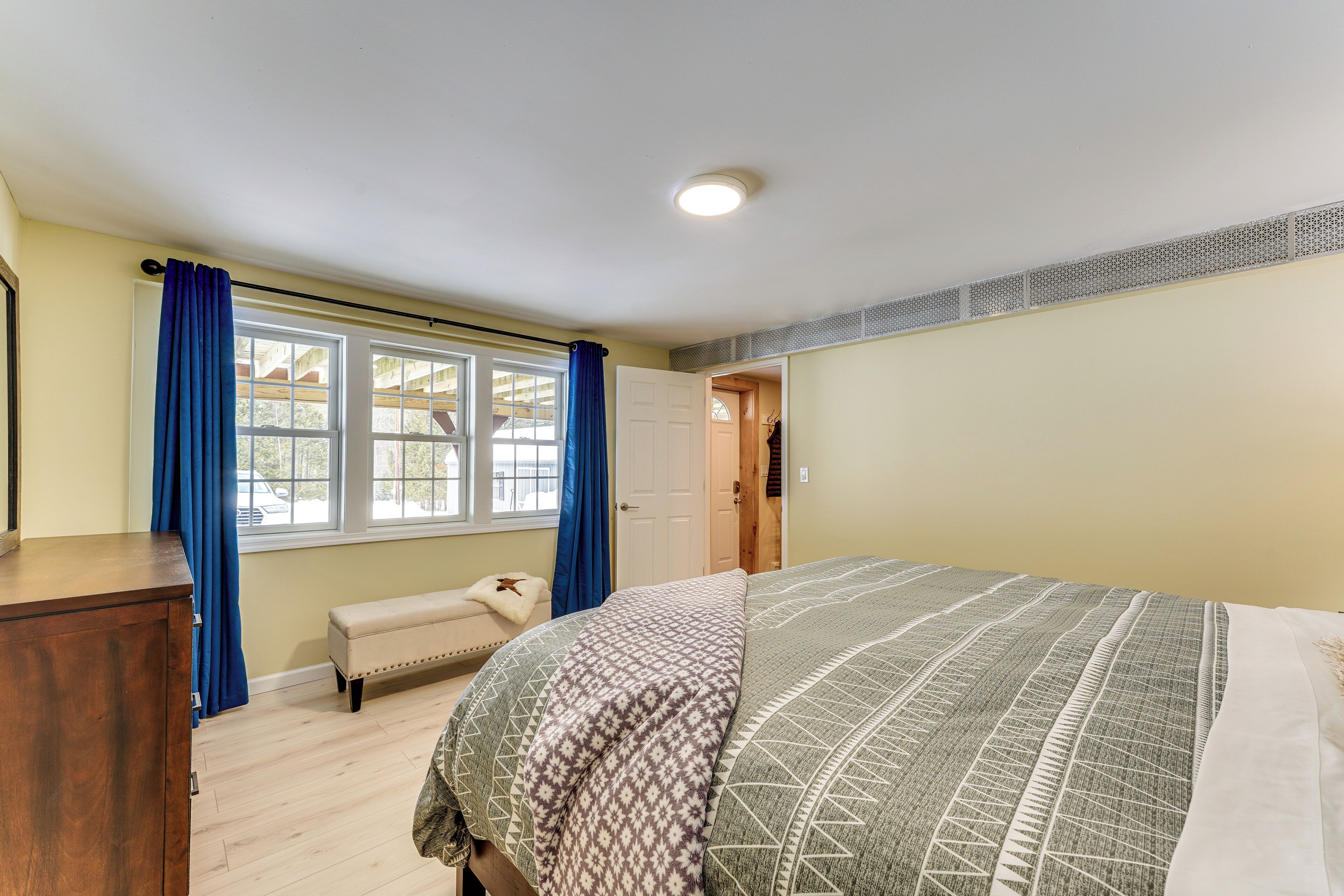 Un dormitorio con cama, cómoda de madera, banco y ventana con cortinas azules.