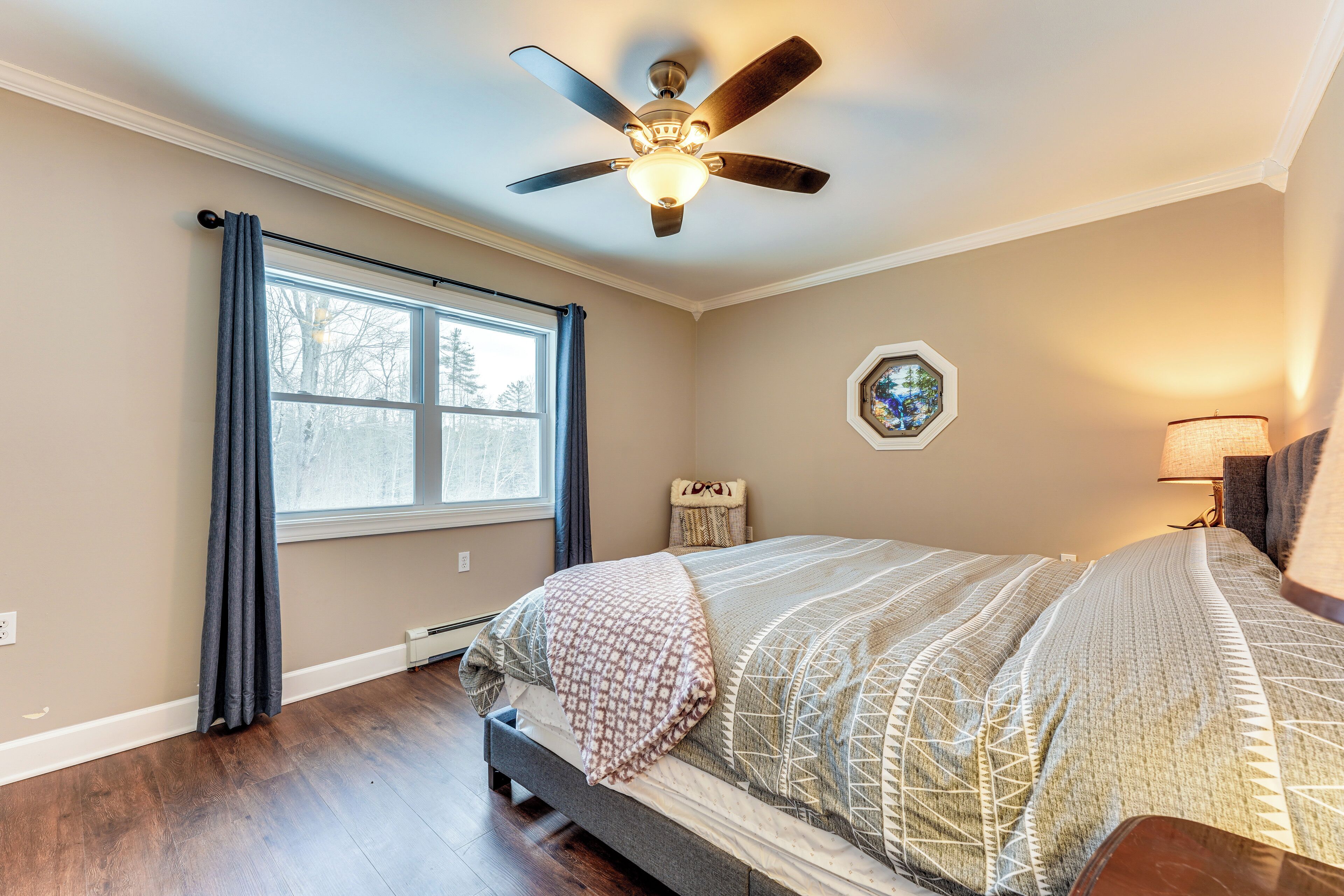 Une chambre à coucher avec un lit, un ventilateur de plafond, une fenêtre avec des rideaux, une table de chevet et une lampe.