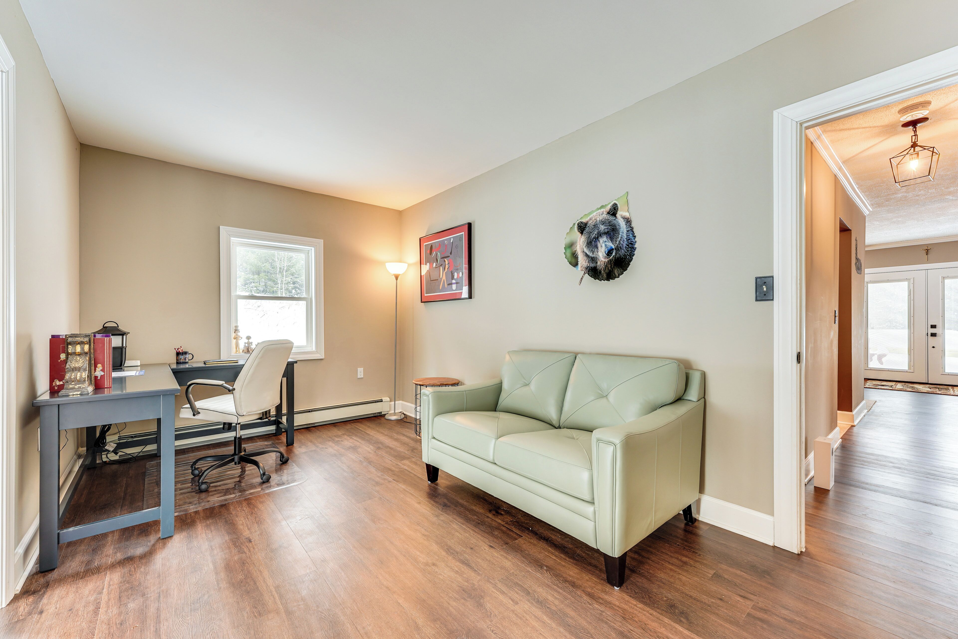 A room with a desk, chair, and a light green sofa. There is a window, a wall-mounted lamp, and a bear head decoration.