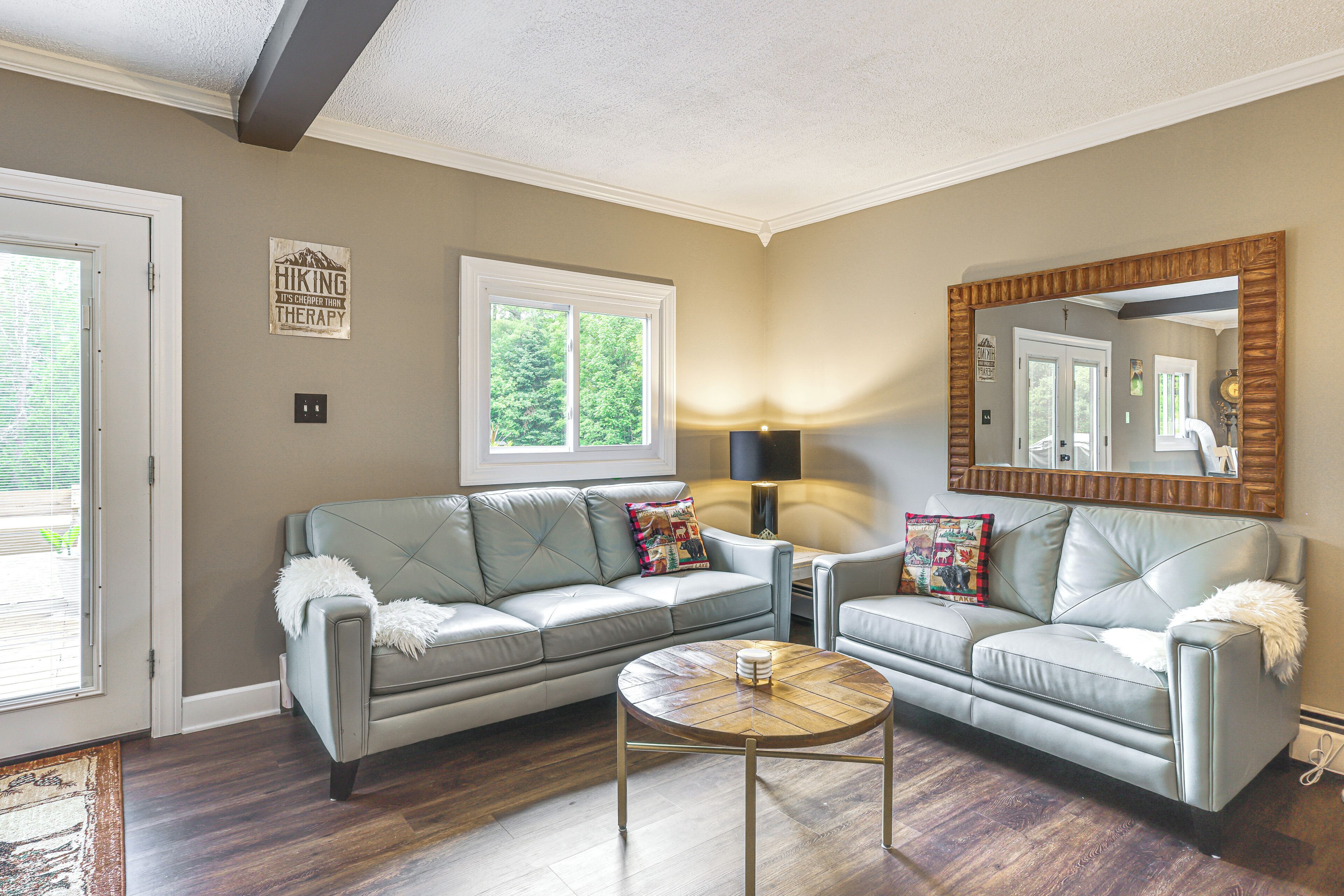 A living room with two grey leather sofas, a round coffee table, a large mirror, and a window with a view of trees.