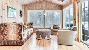 A cozy room with wooden walls and floors, featuring a large window, a sofa, a chair, and a round ottoman.