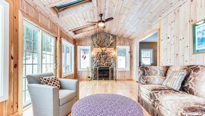 A cozy cabin interior with wooden walls, a stone fireplace, a ceiling fan, and a large window.