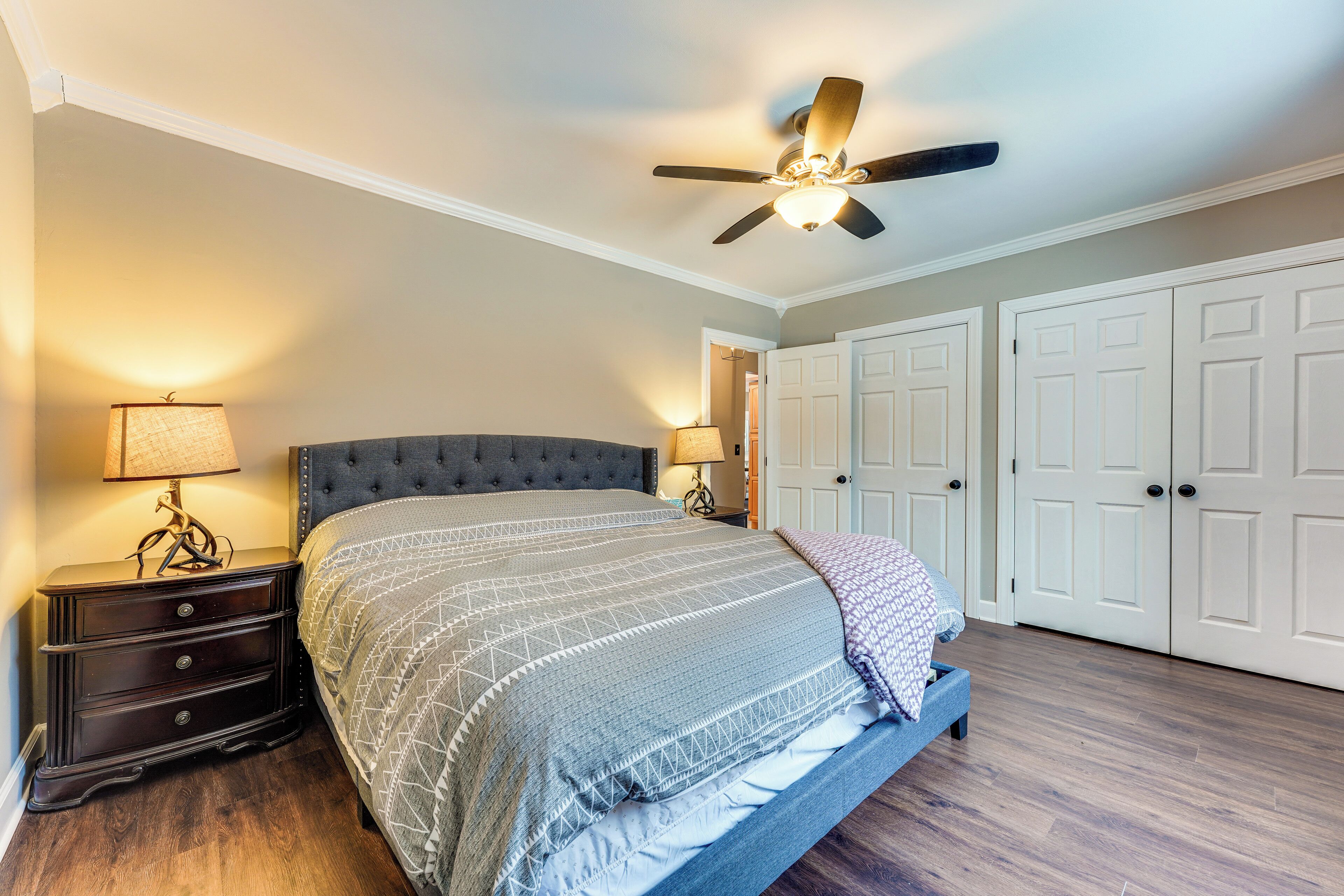 A bedroom with a tufted headboard bed, a ceiling fan, and a nightstand with a lamp.