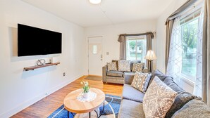 A modern living room with a flat-screen TV mounted on the wall, a wooden coffee table with a vase, a grey sofa with patterned cushions, and a large window with curtains. - Niles Vacation Rental Near St Joseph River! (Niles)