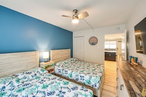 A bedroom with two beds, a ceiling fan, a nightstand, and a kitchen visible through an open door.