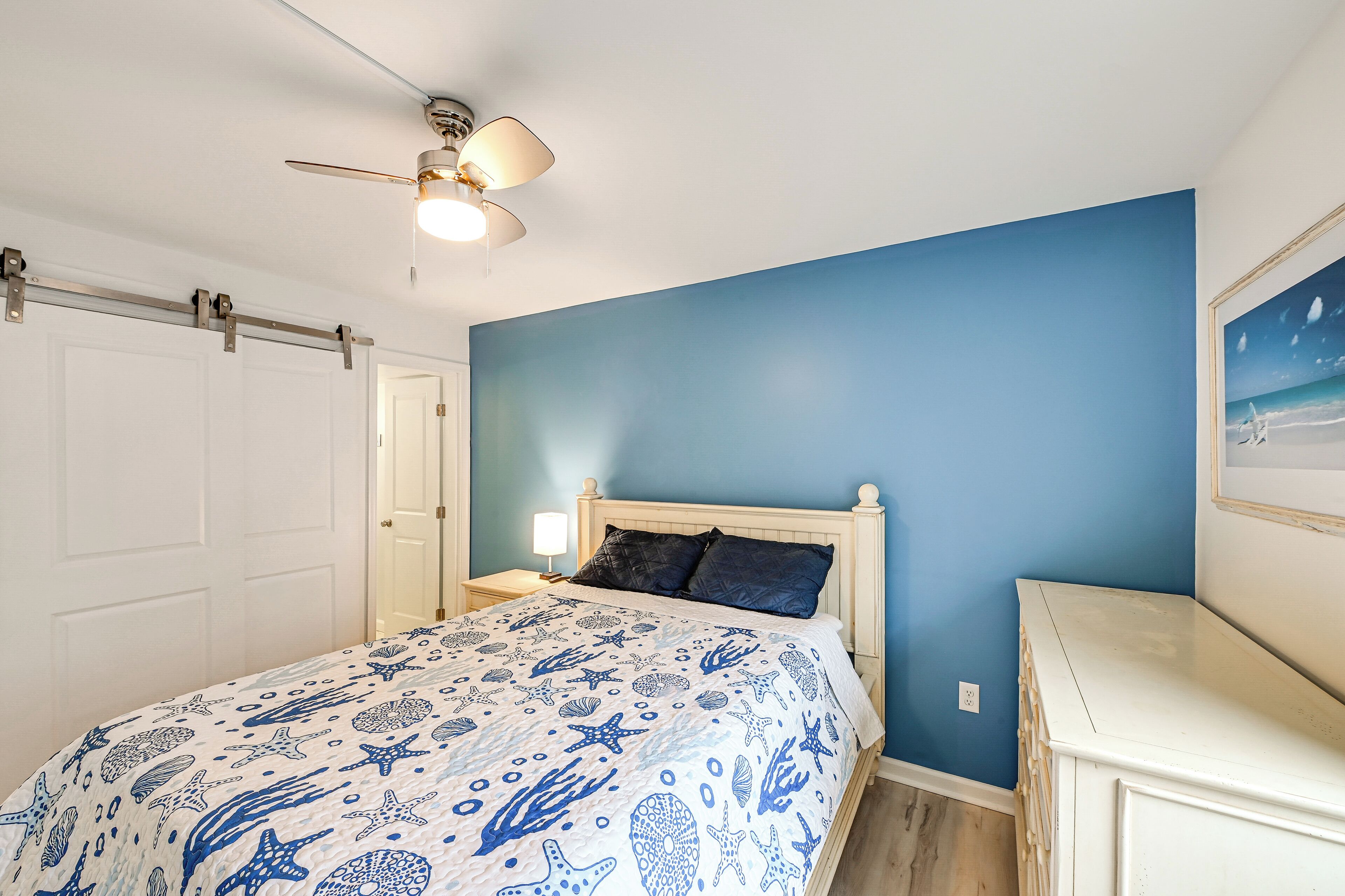 Une chambre à coucher comprenant un lit, une table de chevet, un ventilateur de plafond et un mur orné d’un tableau encadré.