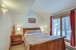 Un dormitorio con cama de madera, una mesita de noche con lámpara, una ventana con cortinas y un cuadro enmarcado en la pared.