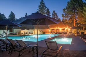 A swimming pool area with lounge chairs and umbrellas, surrounded by trees and a building with outdoor seating. - Pool & River Access: Mt Bachelor Condo w/ Patio! (Bend)