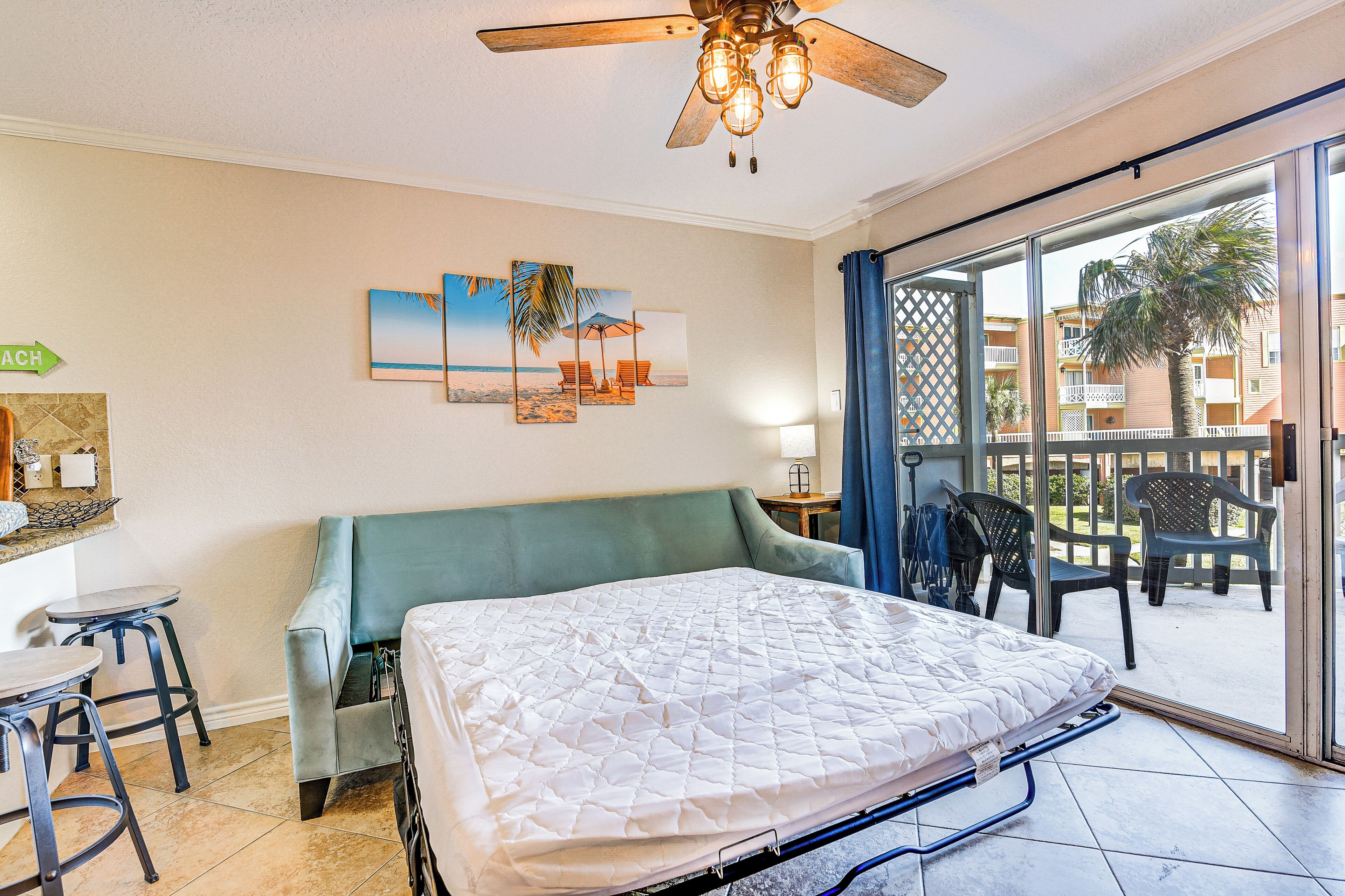 A hotel room with a bed, a ceiling fan, a view of the outside balcony, and artwork on the wall.
