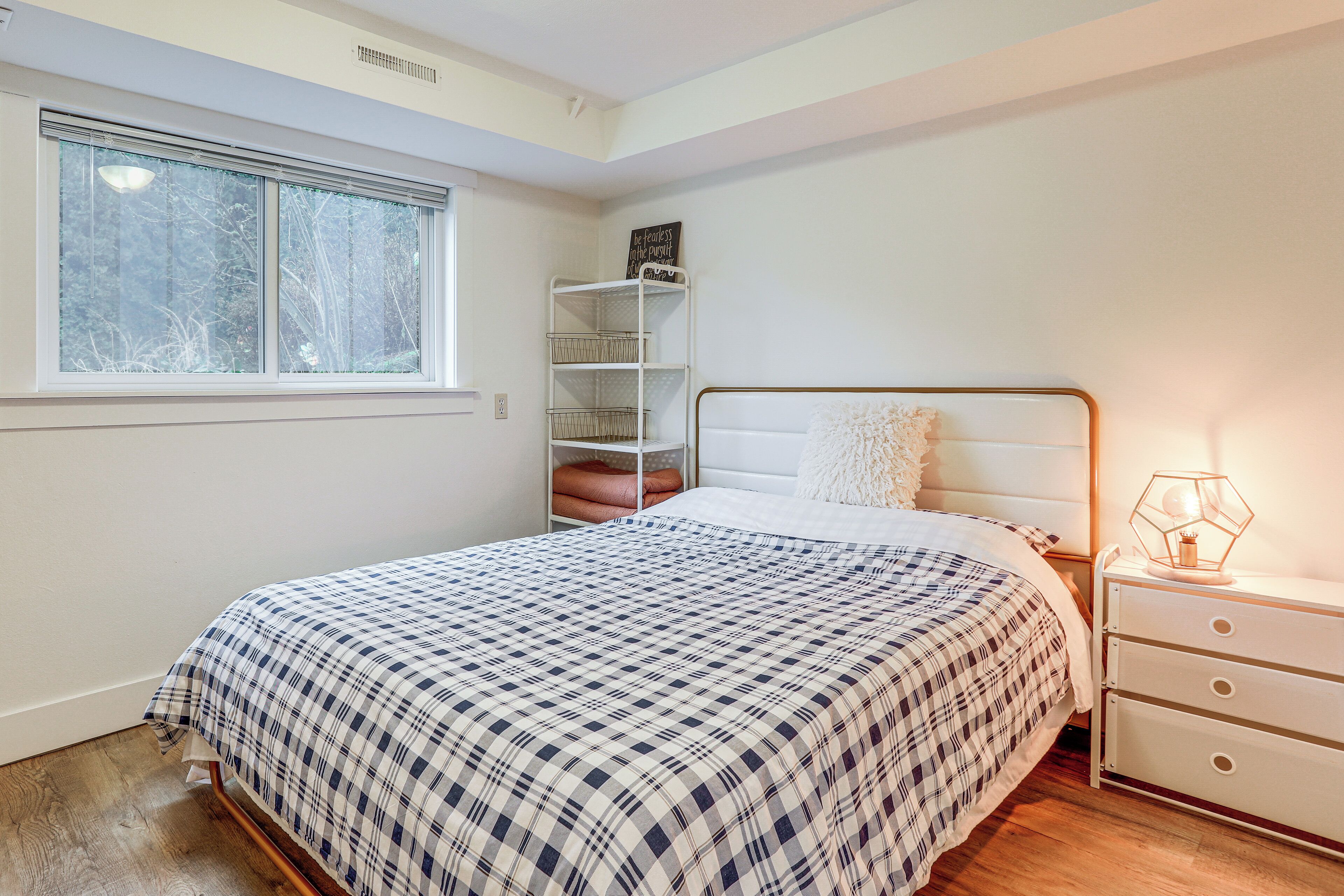Un dormitorio con cama, mesita de noche, lámpara, ventana y estante con cestas.