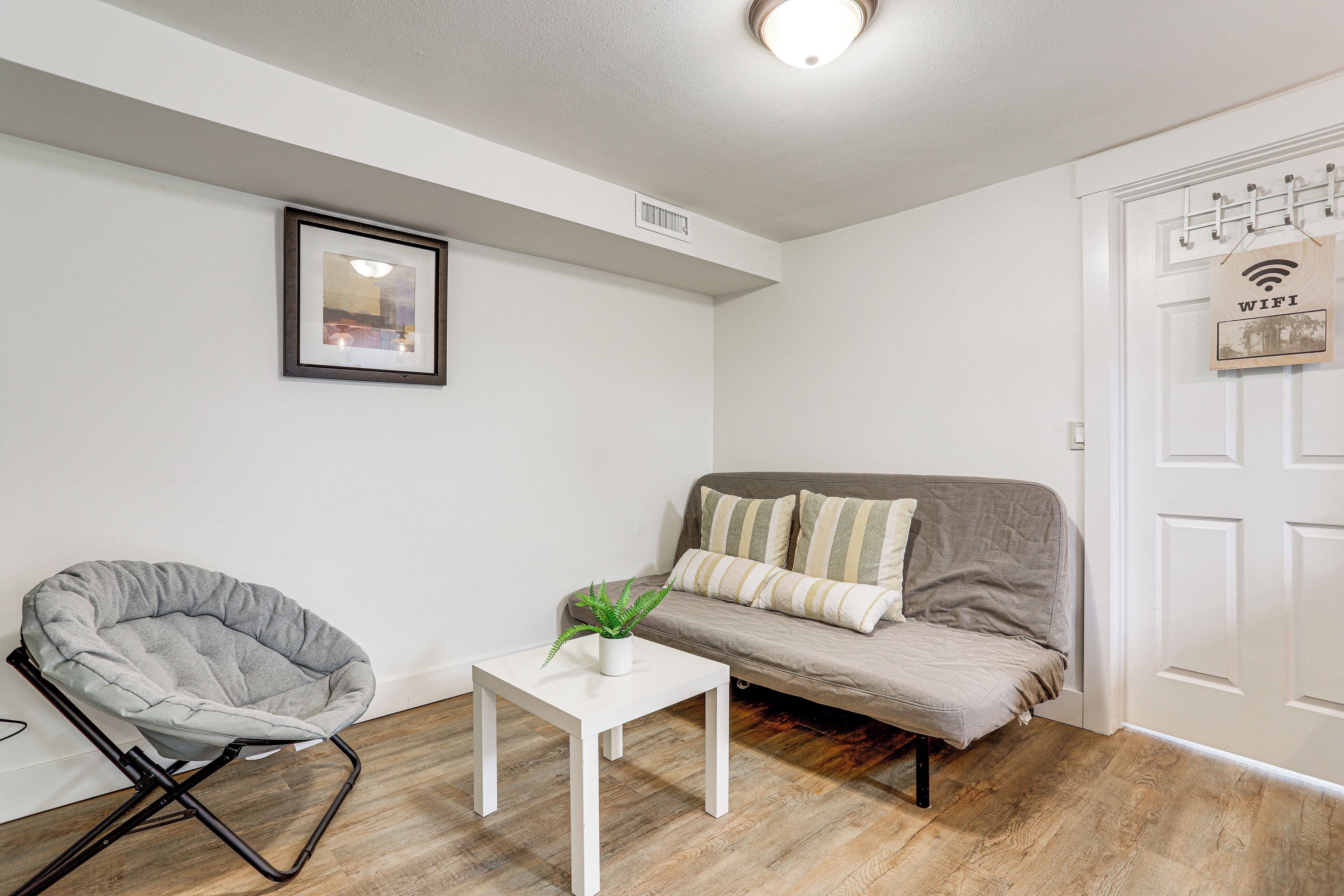 A living room with a grey bean bag chair, a white coffee table with a plant, a grey sofa with striped pillows, and a white door with a 'WiFi' sign.