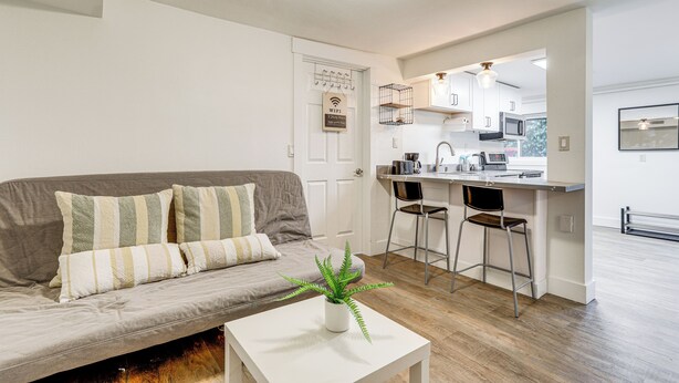 A modern living room with a grey sofa, striped cushions, a white coffee table with a plant, and a kitchen area with a bar stool and a microwave. - Walk to Beach & Lake Access: Cozy Redmond Apt! (Redmond)