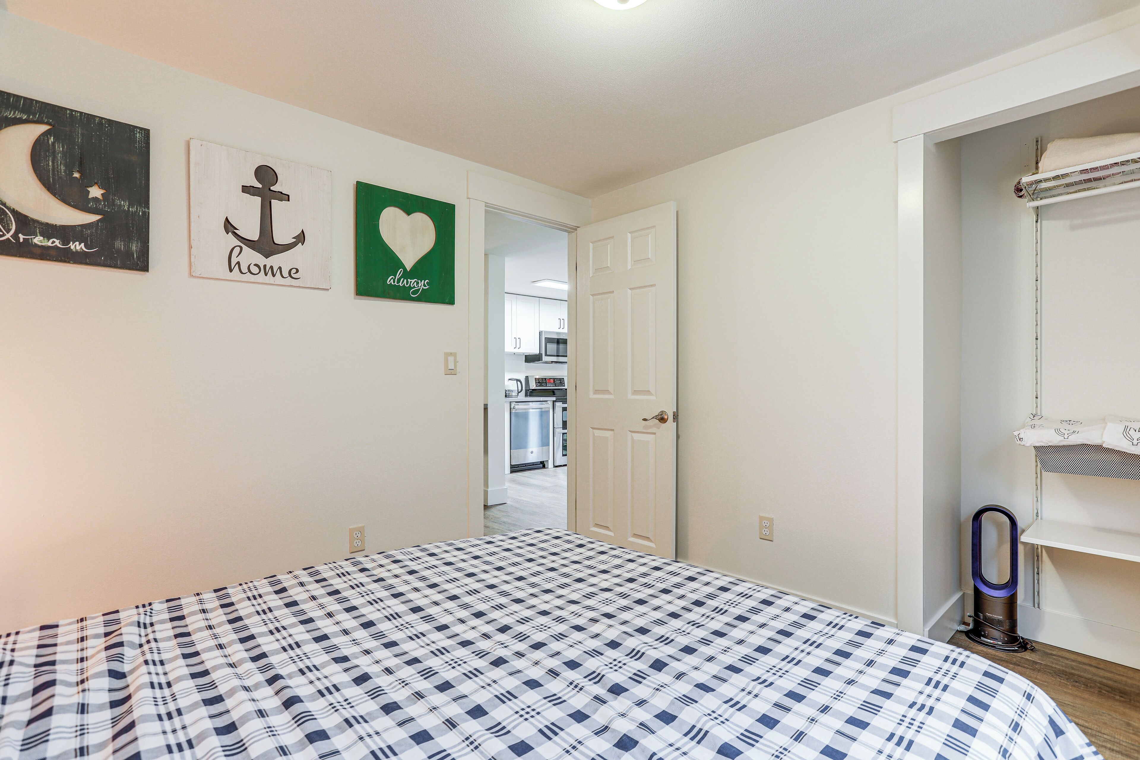 A bedroom with a checkered bedspread, a door leading to a kitchen, and wall art depicting an anchor, a heart, and a moon.
