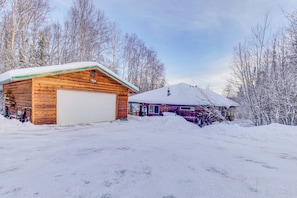 Una cabaña de madera con garaje y techo cubierto de nieve, rodeada de árboles y un cielo despejado.