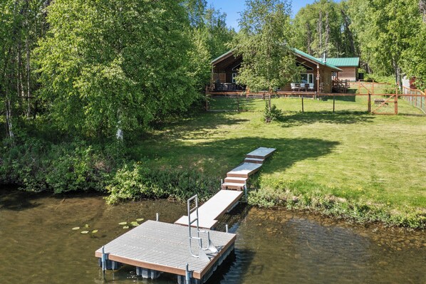 Un muelle de madera se adentra en un río tranquilo, rodeado de abundante vegetación y una cabaña con techo verde.