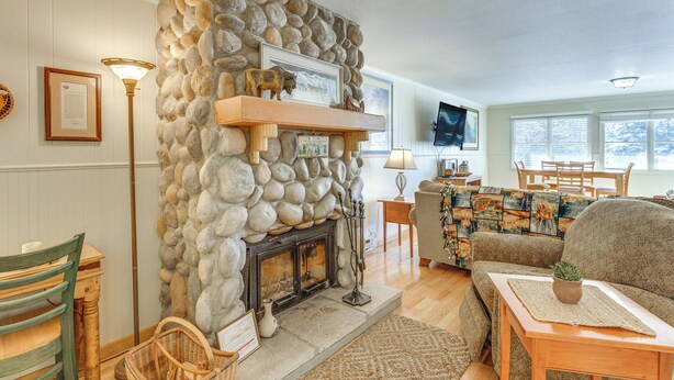 A cozy living room with a stone fireplace, a television mounted on the wall, and a comfortable seating area with a sofa and chairs. - Main Street Red River Condo w/ Fireplace & Views! (Red River)