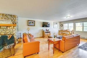 A living room with a stone wall, orange sofas, a wooden dining table, and a TV mounted on the wall. - Main Street Red River Condo w/ Fireplace & Views! (Red River)