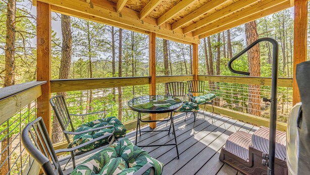 A wooden deck with a glass table and chairs, surrounded by trees. - Ruidoso Cabin Rental w/ Hot Tub: Near Midtown! (Ruidoso)