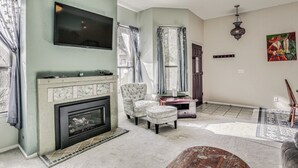 A living room with a fireplace, a television mounted on the wall, and a large window with curtains.