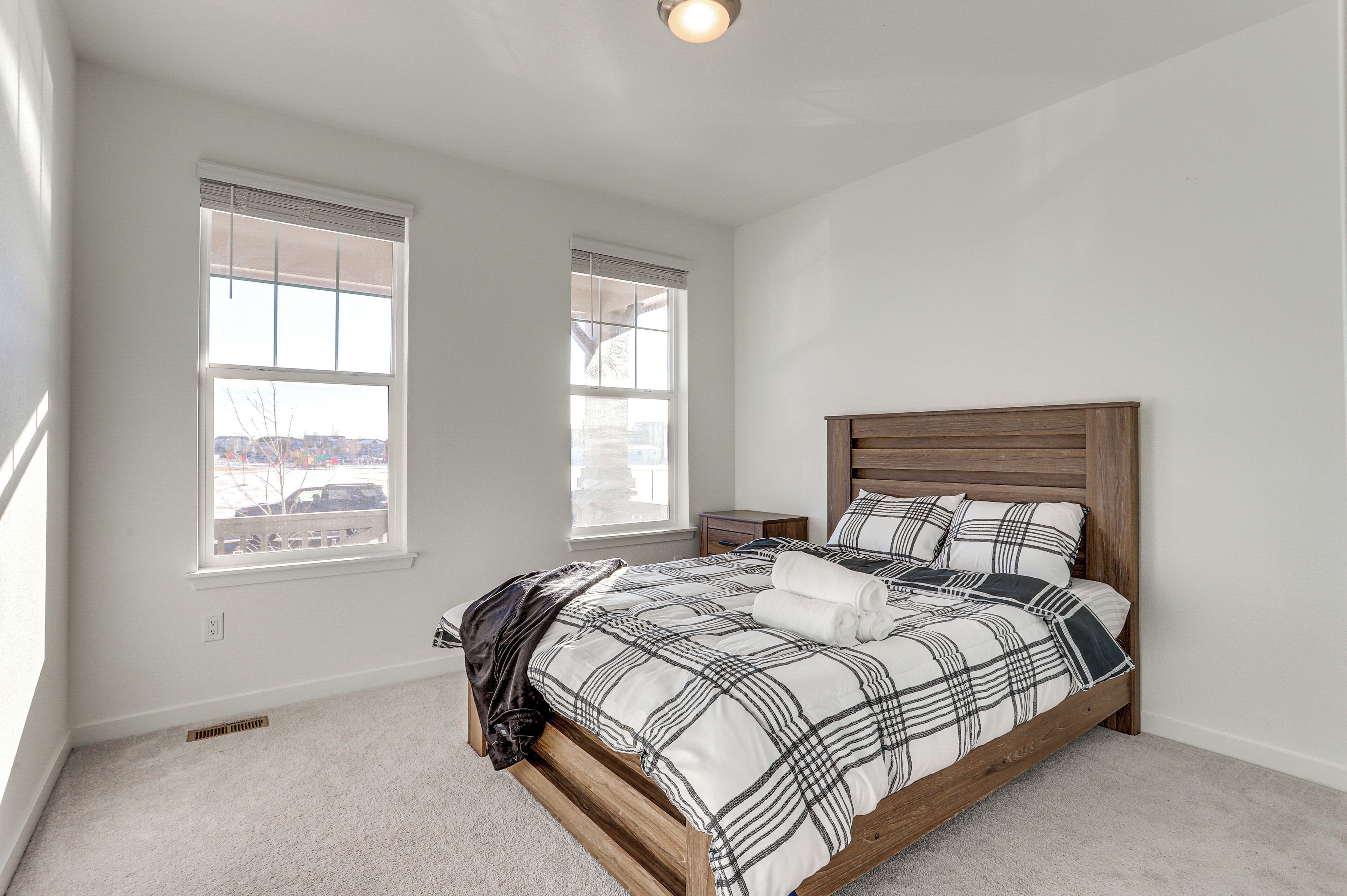 Un dormitorio con estructura de cama de madera, ropa de cama blanca, dos ventanas con persianas, una mesita de noche y una lámpara de techo.