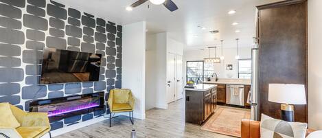 A modern living room with a stone-patterned wall, a fireplace, a ceiling fan, and a view into the kitchen.