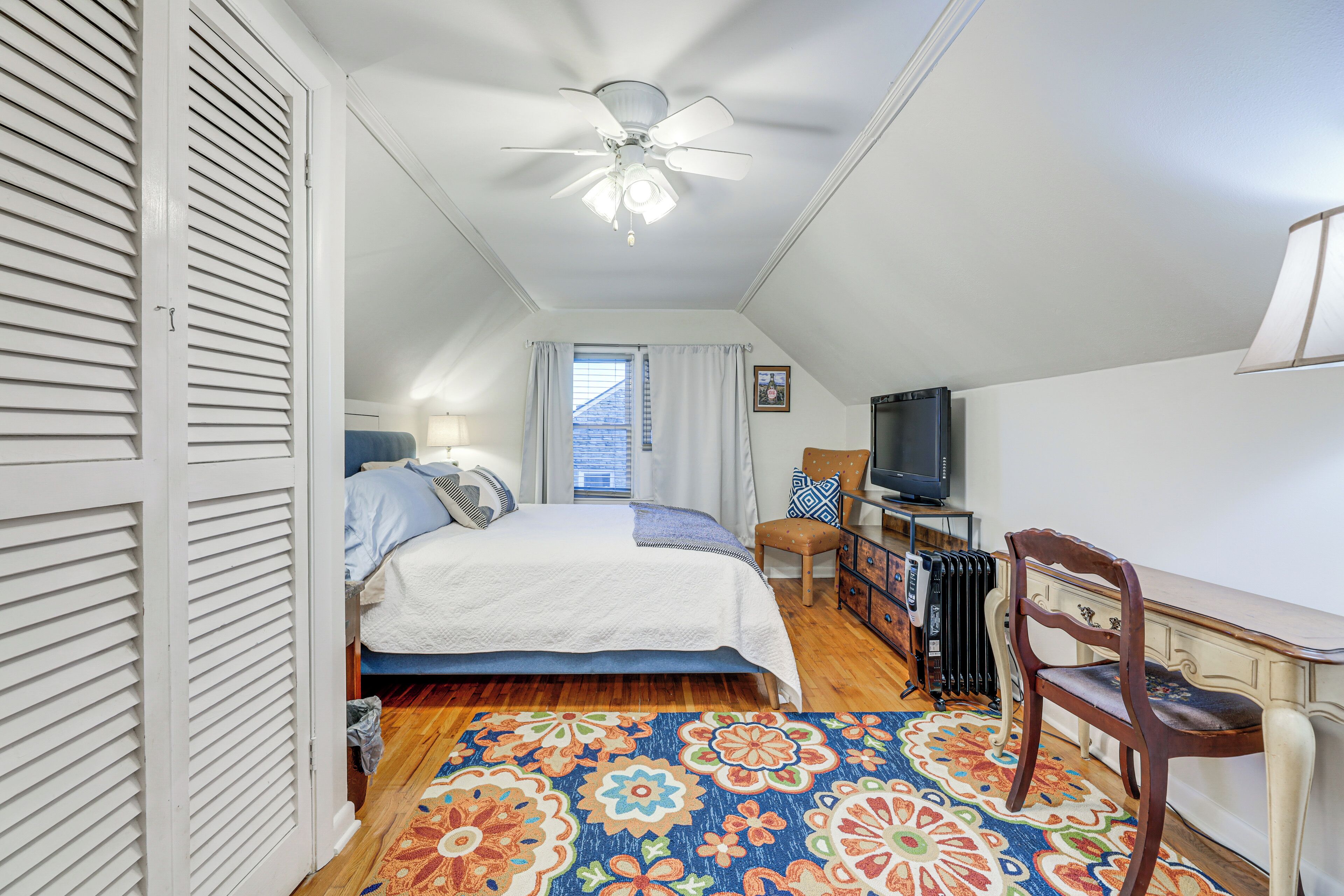 A bedroom with a bed, a desk, a chair, a television, and a ceiling fan.