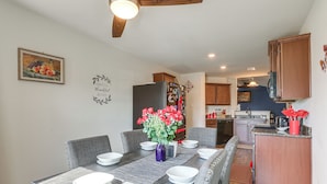Un comedor con una mesa larga dispuesta para seis personas, una cocina al fondo y una pared decorativa con una pintura de frutas y un letrero que dice 'Grateful Blessed'.