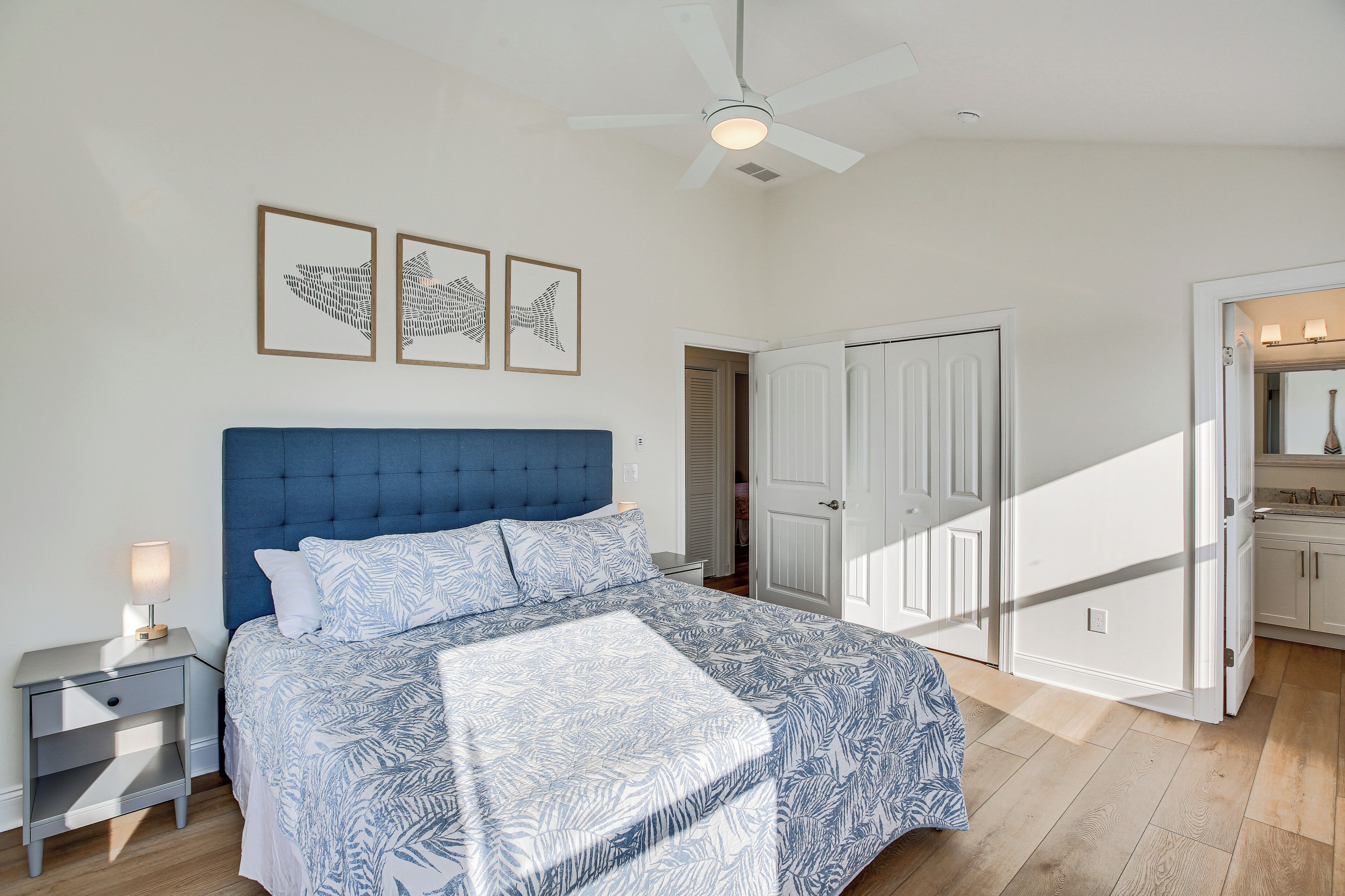 A bedroom with a blue headboard, a bed with patterned bedding, a nightstand with a lamp, and framed artwork on the wall.
