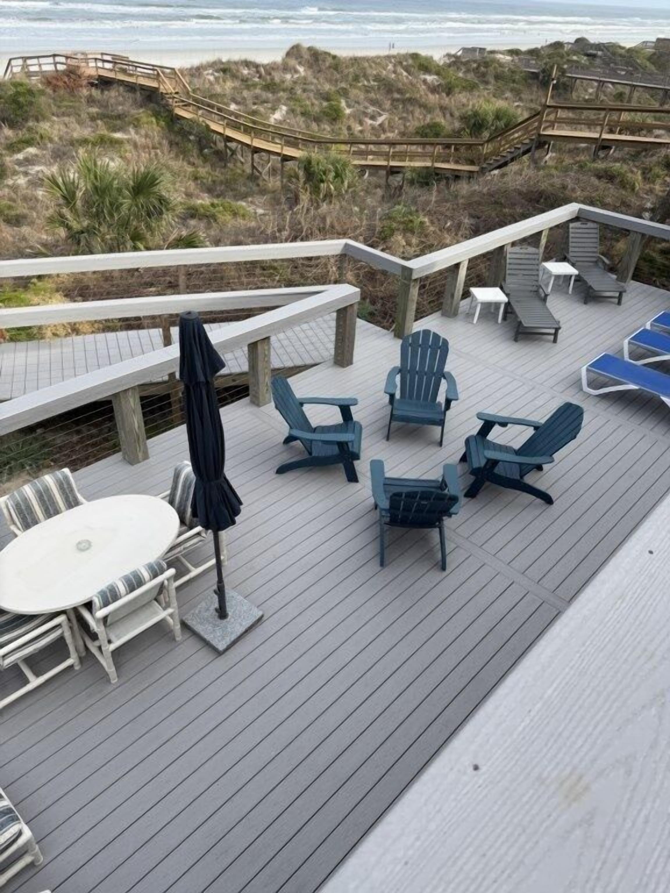 A deck with outdoor seating, including chairs and a table, overlooking a beach and dunes.