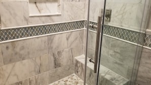 Une salle de bain carrelée de marbre, dotée d’une douche en verre et d’une petite étagère.