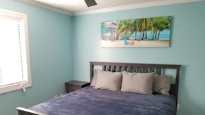 Une chambre à coucher avec un lit, une table de chevet, une fenêtre avec des stores et un tableau représentant une scène de plage.