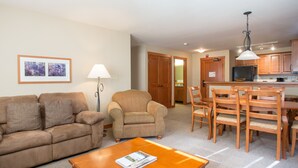 A living room with a sofa, armchair, and coffee table, a dining area with wooden chairs, and a kitchen with wooden cabinets and a refrigerator. - 1202 - Two Bedroom Standard Eagle Springs West 2 Condo by RedAwning (Solitude)