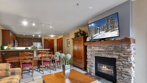 Ein Wohnzimmer mit Kamin aus Stein, eine Essecke mit Holzmöbeln und eine Küche im Hintergrund.