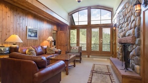 A cozy living room with a leather sofa, wooden walls, a stone fireplace, and a large window.